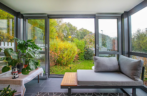 Große Schiebetüren im Maul Wintergarten mit Aluminiumrahmen und Wärmeschutzverglasung öffnen den Blick ins Grün, drinnen Sitzbank und Pflanzen.