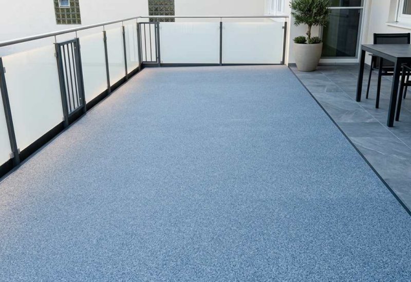Der blau-graue Balkonbelag mit feiner Struktur und die klaren Glasgeländer prägen die Anbaubalkon-Optik und ergänzen die moderne Fassadenlinie.