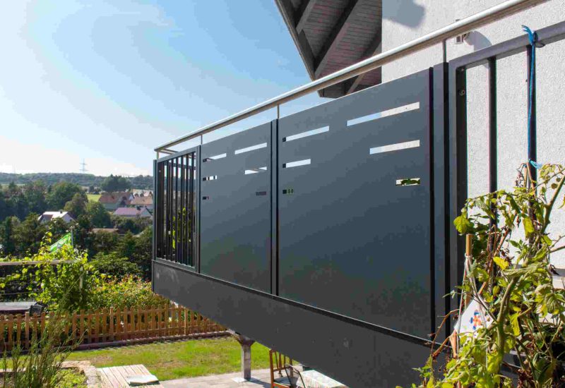Dunkelgraues Glasgeländer am Anbaubalkon mit vertikalen Stahlstreben, klaren Glasfüllungen und Blick ins Grüne bei Tageslicht.