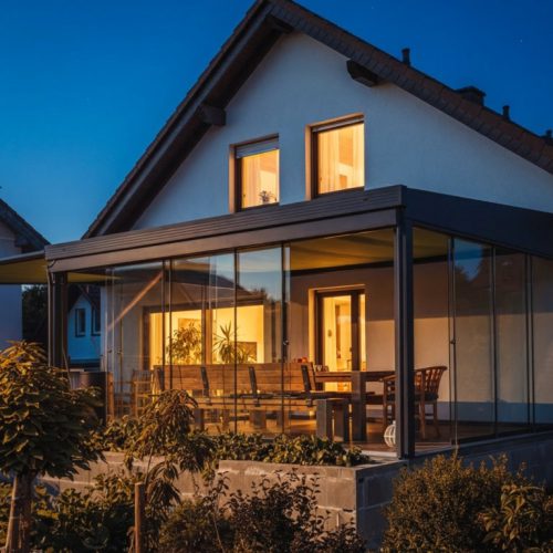 Sommergarten in Maul bei Nacht mit verglaster Überdachung, warmem Licht im Innenraum und Holzterrasse am Haus unter blauem Himmel für Licht, Raum und Qualität.