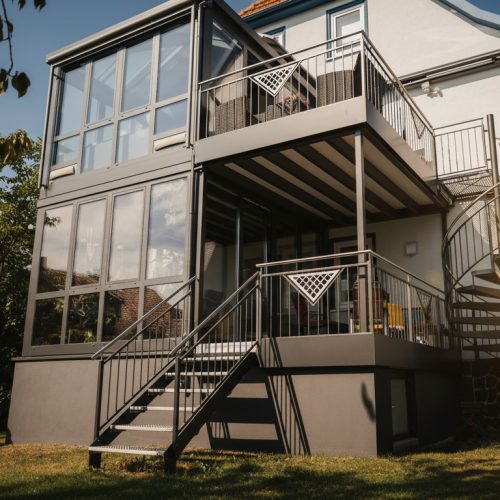 Verglaster Wintergarten mit Anbaubalkon am Haus, schwarze Rahmung und mehrstufige Treppe sorgen für mehr Licht und Raumgefühl im Alltag.