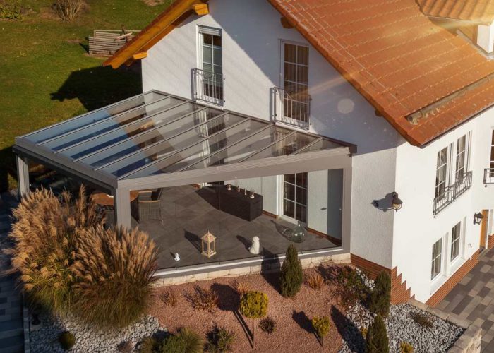 Ein Maul Wintergarten mit großen Glasflächen und aluminiumfarbenem Rahmen über Terrasse und Sitzbereich, daneben Haus und Garten mit weichem Grün.