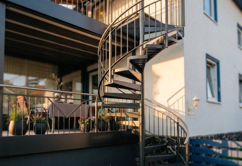 Geschwungene Treppe mit metallischem Geländer verbindet Ebenen am Anbaubalkon, im Hintergrund ruhige Architektur und dezente Begrünung im Außenbereich.