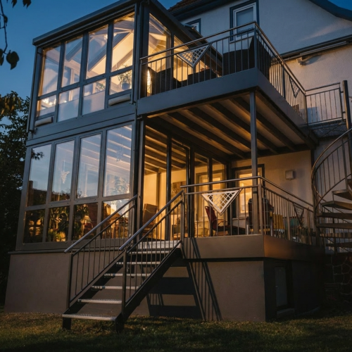 Moderne Wintergarten-Architektur mit Aluminiumkonstruktion und großen Glasflächen, angrenzendem Anbaubalkon und warmem Licht bei Dämmerung.